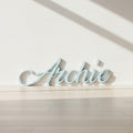 Personalized cursive name sign reading 'Archie' in a pale light blue color, displayed on a gray concrete floor.