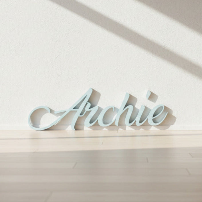 Personalized cursive name sign reading 'Archie' in a pale light blue color, displayed on a gray concrete floor.